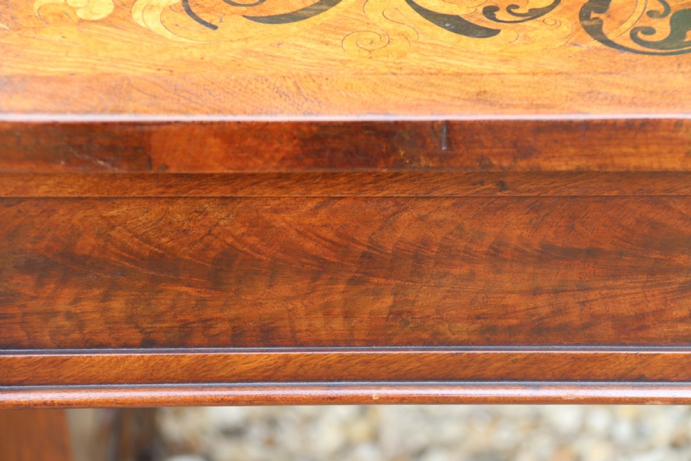 An early 19th-century flame mahogany library table featuring floral marquetry top borders, two frieze drawers with brass knobs, and carved trestle legs connected by a turned stretcher. View XVII