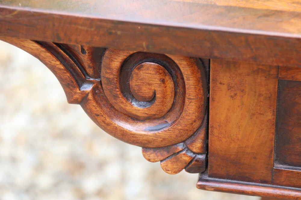 An early 19th-century flame mahogany library table featuring floral marquetry top borders, two frieze drawers with brass knobs, and carved trestle legs connected by a turned stretcher. View XVIII