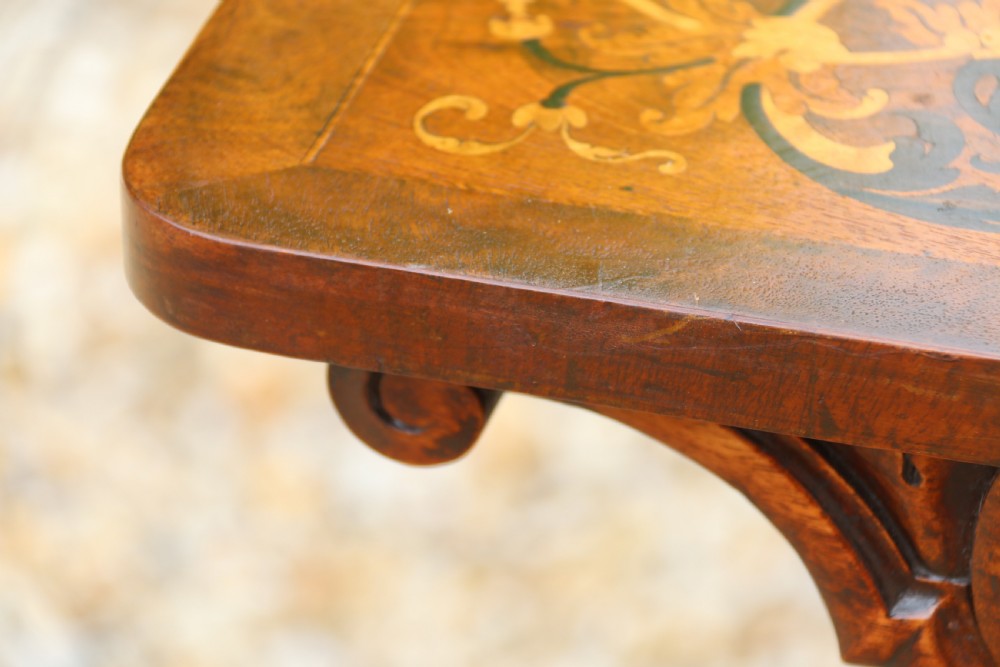 An early 19th-century flame mahogany library table featuring floral marquetry top borders, two frieze drawers with brass knobs, and carved trestle legs connected by a turned stretcher. View XIX