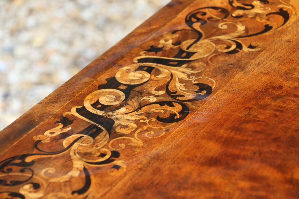 An early 19th-century flame mahogany library table featuring floral marquetry top borders, two frieze drawers with brass knobs, and carved trestle legs connected by a turned stretcher. View XXVIII
