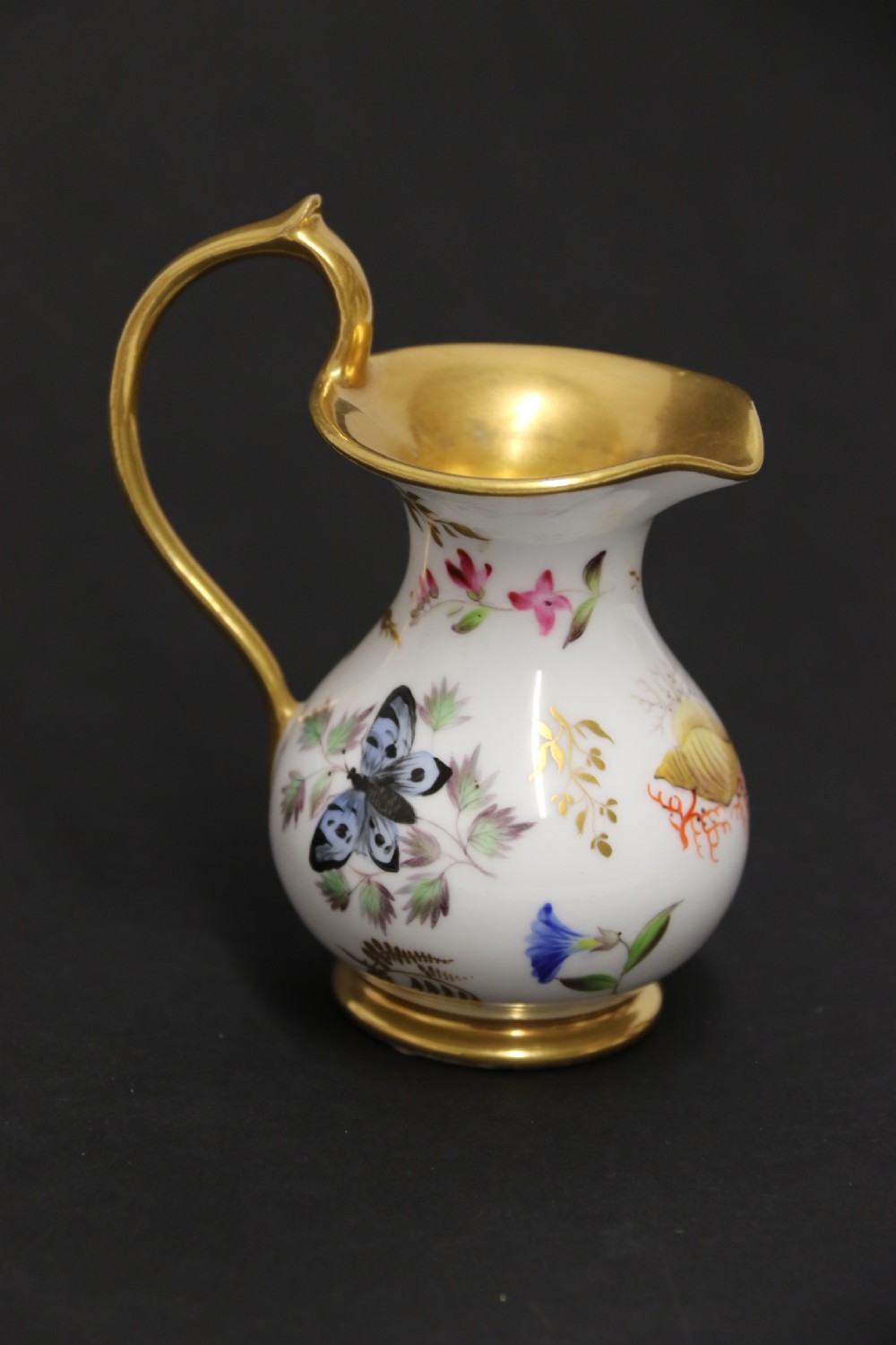A small white hard-paste porcelain ewer with a high looped gilded handle, flared gold interior rim, and hand-painted motifs including a butterfly, seashells, and botanical leaves.