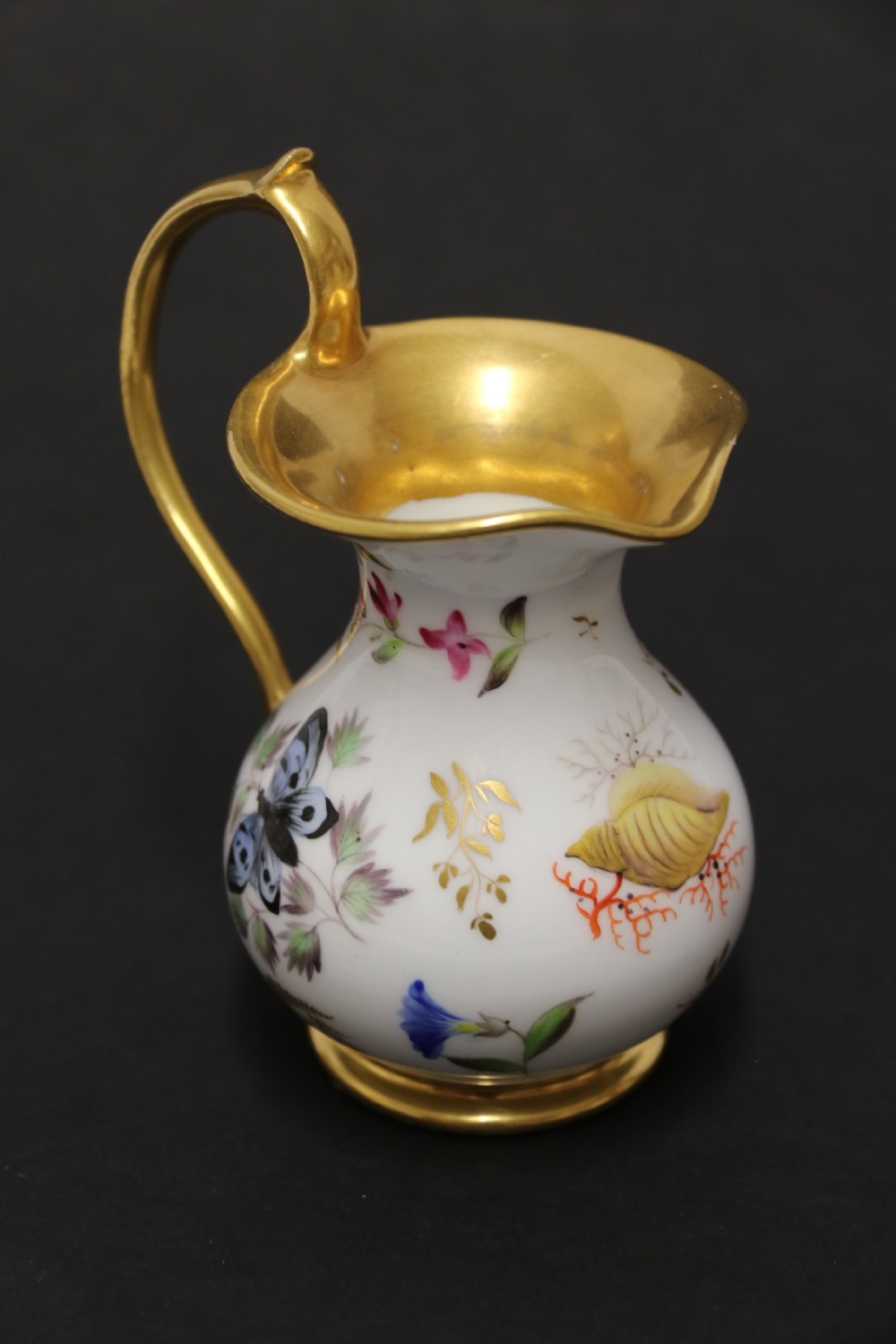 A small white hard-paste porcelain ewer with a high looped gilded handle, flared gold interior rim, and hand-painted motifs including a butterfly, seashells, and botanical leaves. View IV