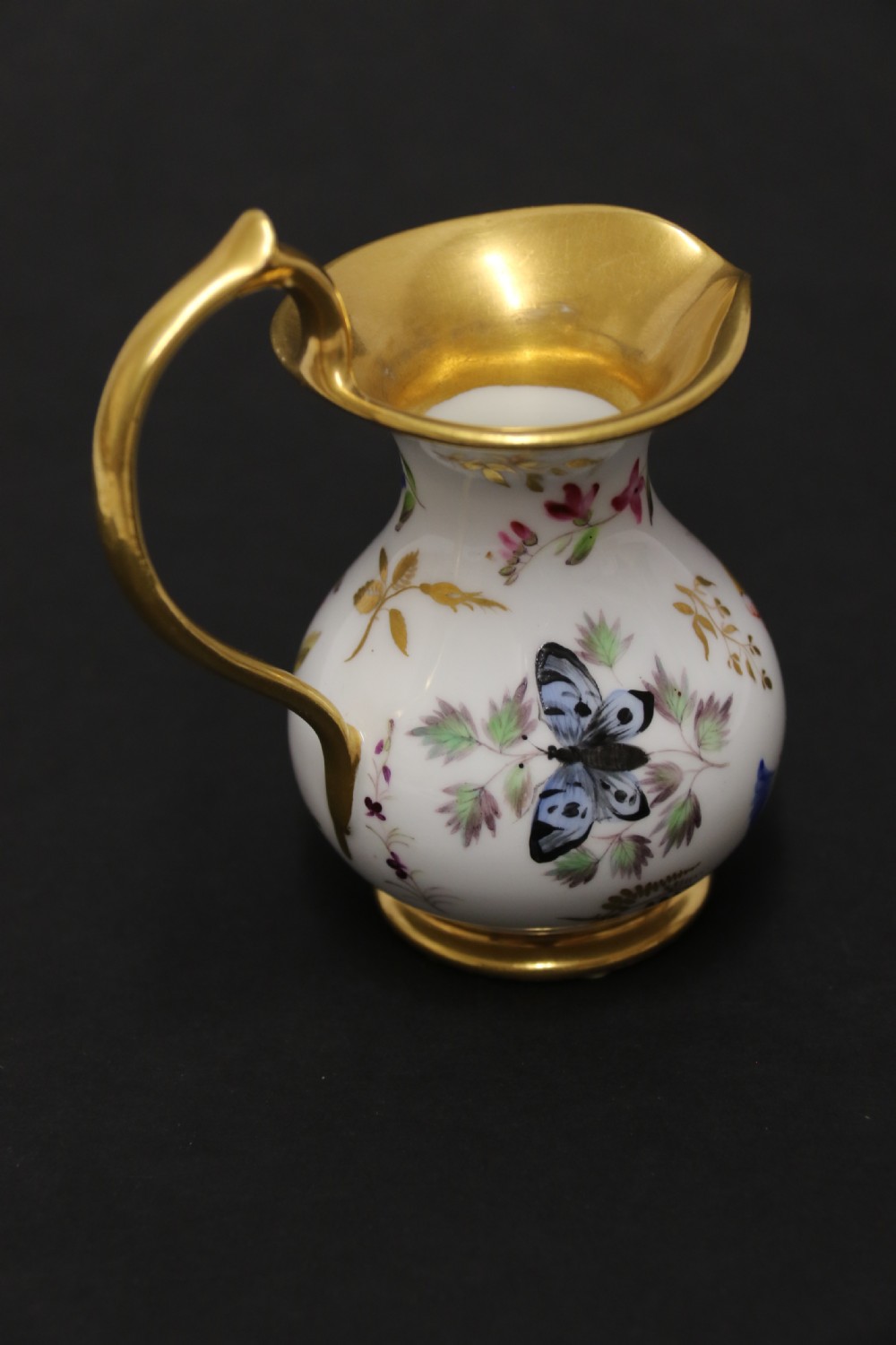 A small white hard-paste porcelain ewer with a high looped gilded handle, flared gold interior rim, and hand-painted motifs including a butterfly, seashells, and botanical leaves. View V