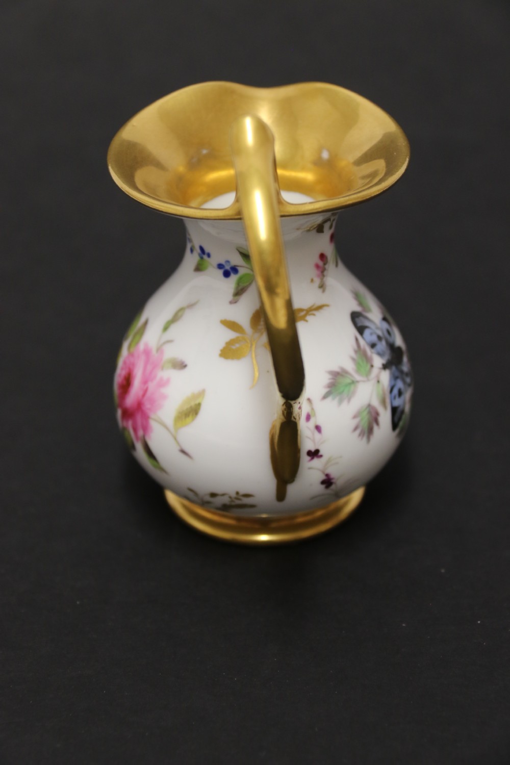 A small white hard-paste porcelain ewer with a high looped gilded handle, flared gold interior rim, and hand-painted motifs including a butterfly, seashells, and botanical leaves. View VI