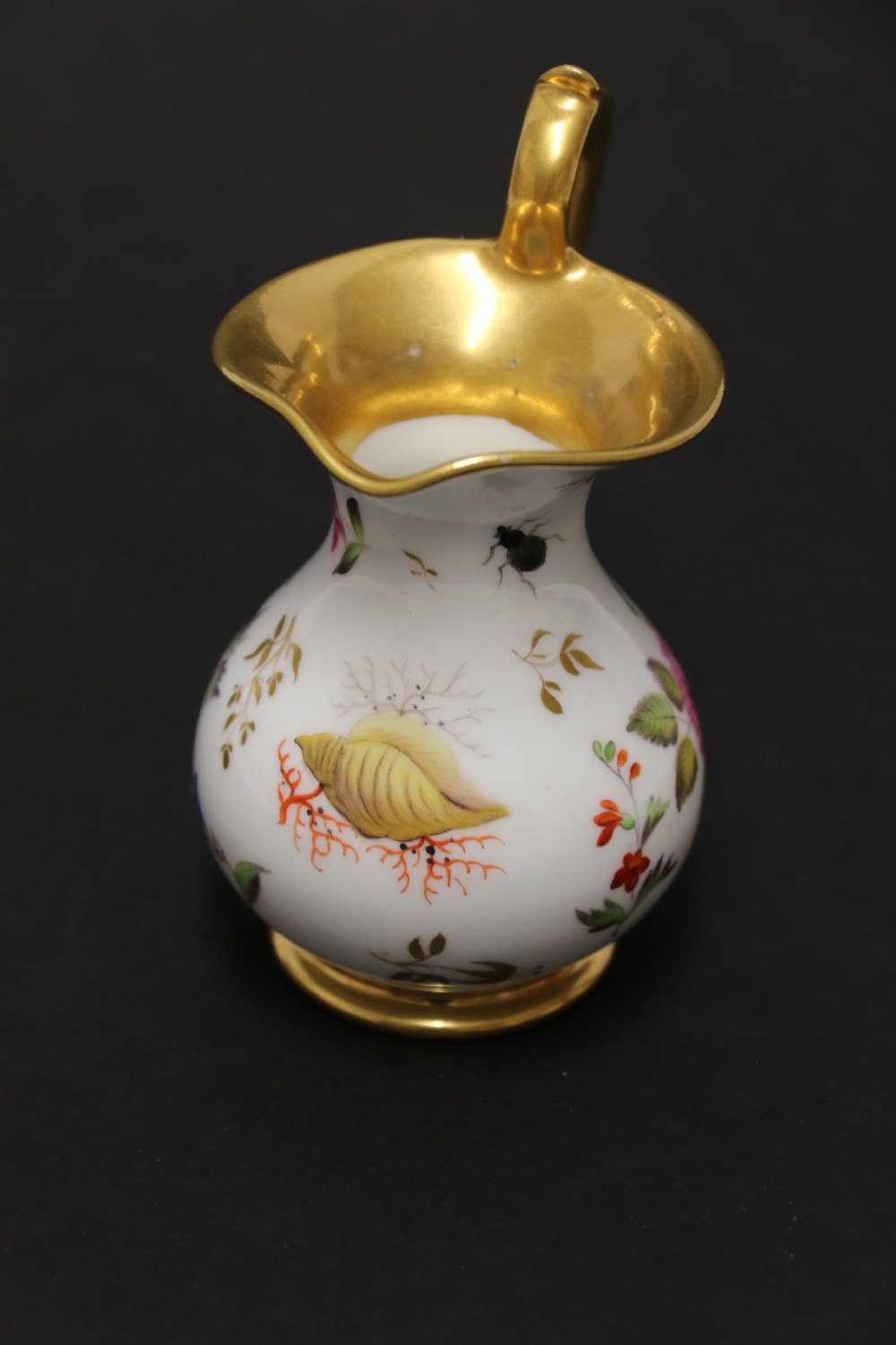 A small white hard-paste porcelain ewer with a high looped gilded handle, flared gold interior rim, and hand-painted motifs including a butterfly, seashells, and botanical leaves. View VII