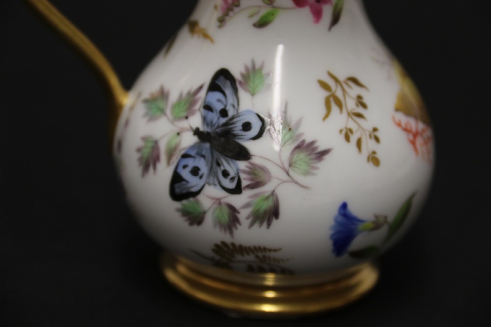 A small white hard-paste porcelain ewer with a high looped gilded handle, flared gold interior rim, and hand-painted motifs including a butterfly, seashells, and botanical leaves. View X