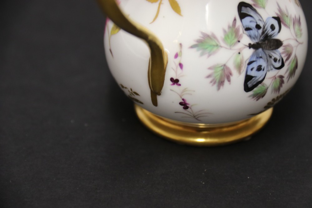 A small white hard-paste porcelain ewer with a high looped gilded handle, flared gold interior rim, and hand-painted motifs including a butterfly, seashells, and botanical leaves. View XI