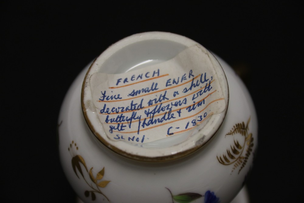 A small white hard-paste porcelain ewer with a high looped gilded handle, flared gold interior rim, and hand-painted motifs including a butterfly, seashells, and botanical leaves. View XII