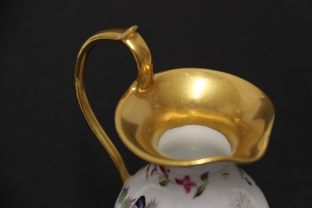 A small white hard-paste porcelain ewer with a high looped gilded handle, flared gold interior rim, and hand-painted motifs including a butterfly, seashells, and botanical leaves. View XIII