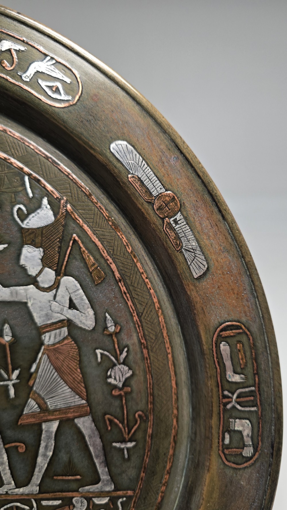 A circular bronze charger plate inlaid with silver and copper depicting ancient Egyptian figures, winged scarabs, and hieroglyphic motifs. View XII