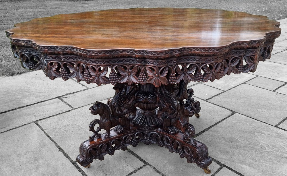 A 19th-century Anglo-Indian carved padauk wood centre table featuring a scalloped top, deeply carved pierced apron of foliage, and a pedestal base supported by carved mythical lion feet with brass castors.