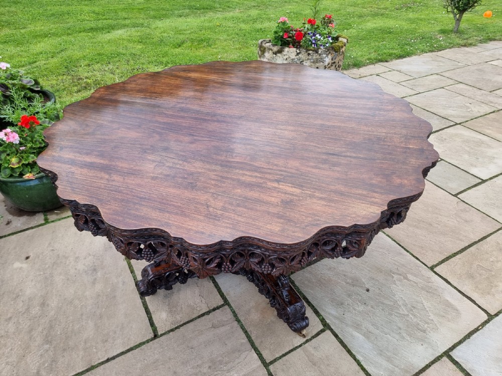 A 19th-century Anglo-Indian carved padauk wood centre table featuring a scalloped top, deeply carved pierced apron of foliage, and a pedestal base supported by carved mythical lion feet with brass castors. View III