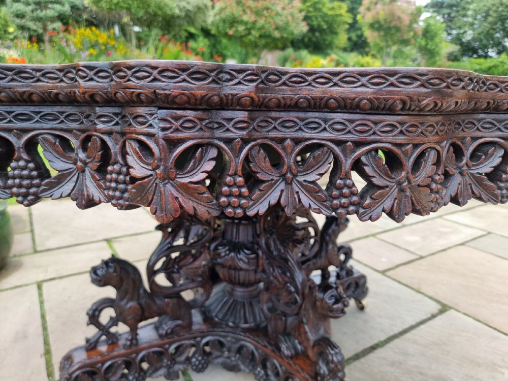 A 19th-century Anglo-Indian carved padauk wood centre table featuring a scalloped top, deeply carved pierced apron of foliage, and a pedestal base supported by carved mythical lion feet with brass castors. View VI