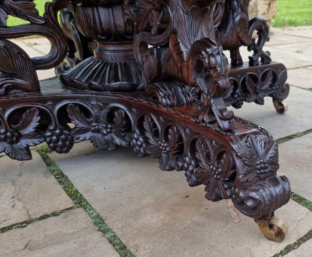 A 19th-century Anglo-Indian carved padauk wood centre table featuring a scalloped top, deeply carved pierced apron of foliage, and a pedestal base supported by carved mythical lion feet with brass castors. View VIII