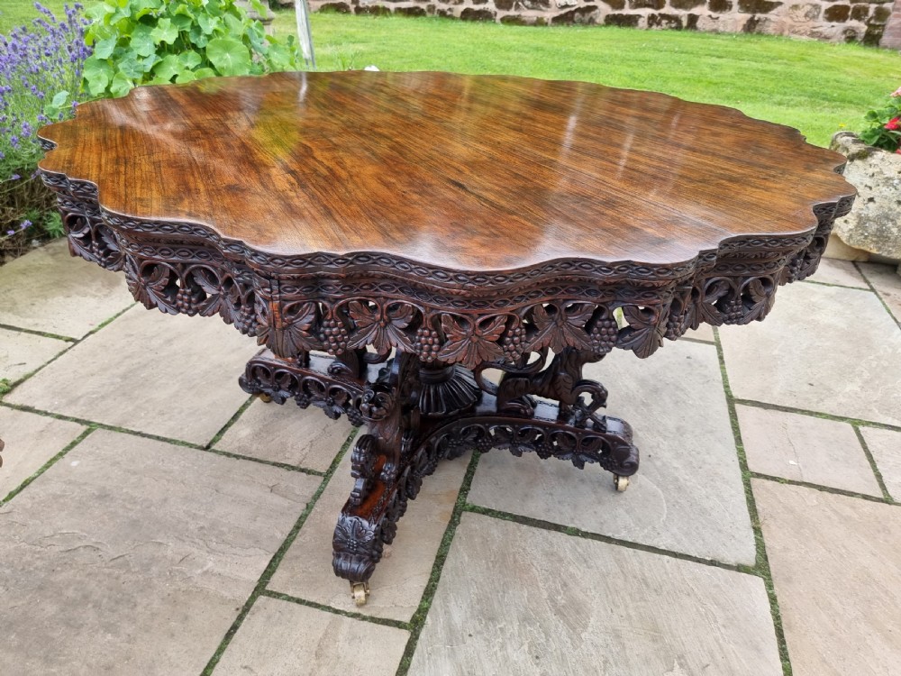A 19th-century Anglo-Indian carved padauk wood centre table featuring a scalloped top, deeply carved pierced apron of foliage, and a pedestal base supported by carved mythical lion feet with brass castors. View IX