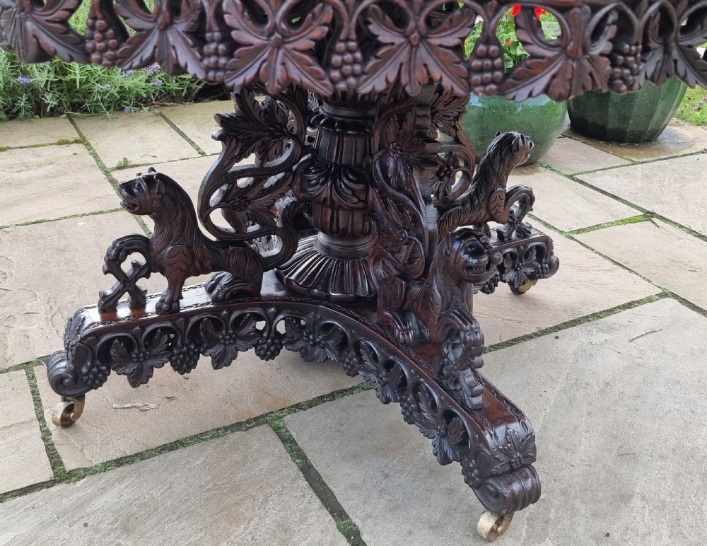 A 19th-century Anglo-Indian carved padauk wood centre table featuring a scalloped top, deeply carved pierced apron of foliage, and a pedestal base supported by carved mythical lion feet with brass castors. View XVI