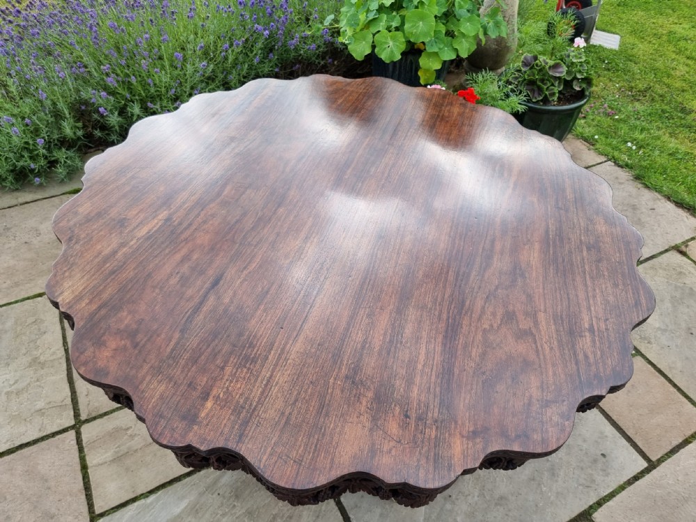 A 19th-century Anglo-Indian carved padauk wood centre table featuring a scalloped top, deeply carved pierced apron of foliage, and a pedestal base supported by carved mythical lion feet with brass castors. View XVII