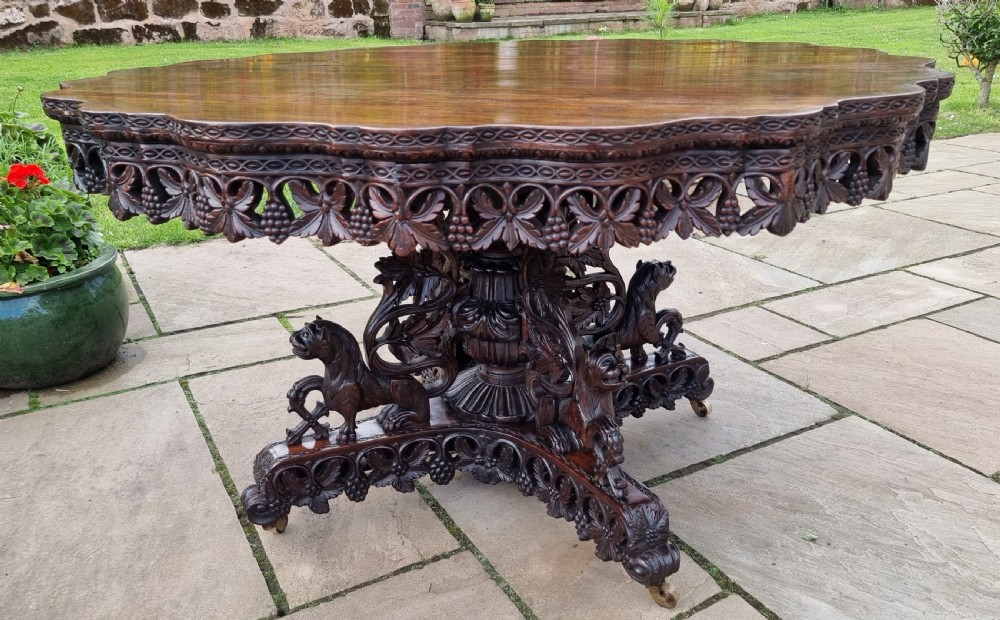 A 19th-century Anglo-Indian carved padauk wood centre table featuring a scalloped top, deeply carved pierced apron of foliage, and a pedestal base supported by carved mythical lion feet with brass castors. View XVIII