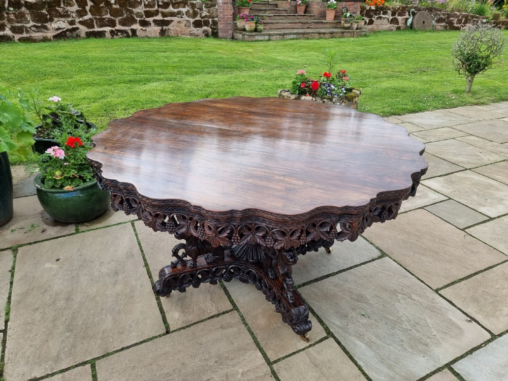 A 19th-century Anglo-Indian carved padauk wood centre table featuring a scalloped top, deeply carved pierced apron of foliage, and a pedestal base supported by carved mythical lion feet with brass castors. View XIX
