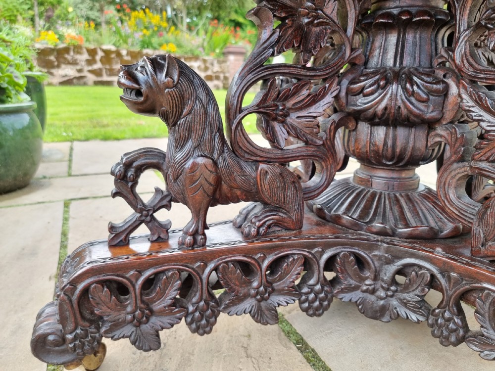 A 19th-century Anglo-Indian carved padauk wood centre table featuring a scalloped top, deeply carved pierced apron of foliage, and a pedestal base supported by carved mythical lion feet with brass castors. View XX