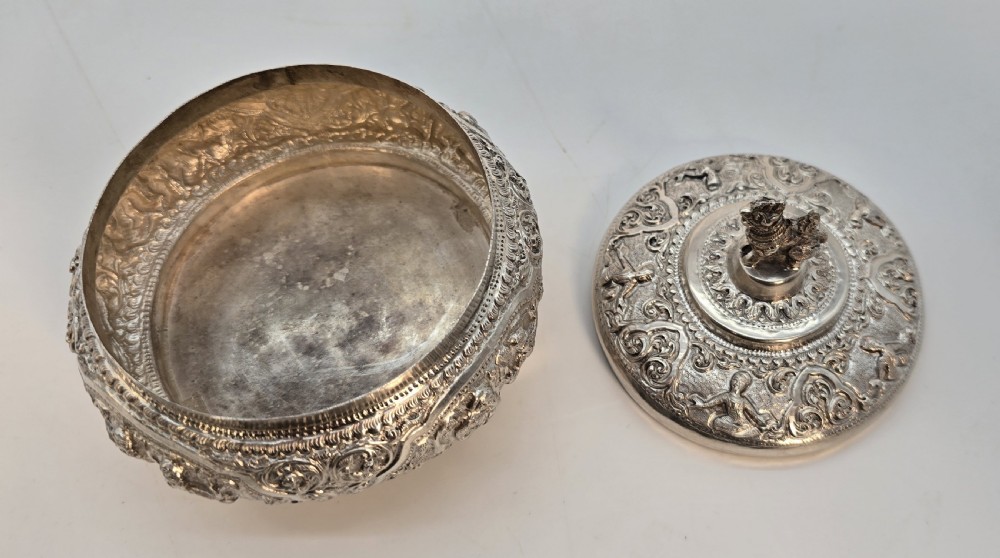 An intricate silver lidded jar featuring raised repousse decorative carving around the body and a small animal finial on top. View XI