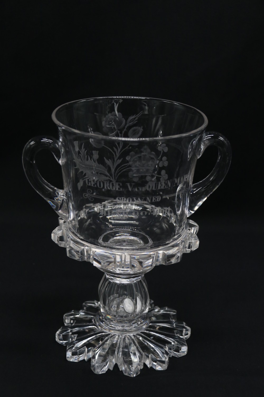A clear glass commemorative goblet with two loop handles, wheel-engraved with text for the 1911 coronation of King George V and Queen Mary, supported by a faceted stem and starburst base.