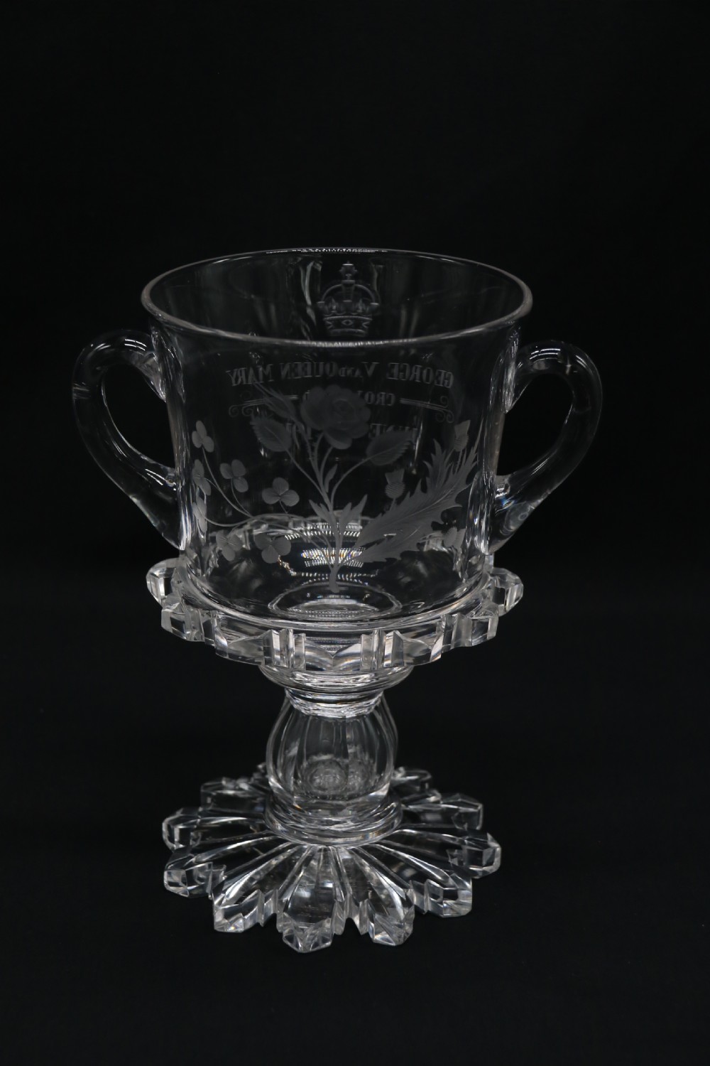 A clear glass commemorative goblet with two loop handles, wheel-engraved with text for the 1911 coronation of King George V and Queen Mary, supported by a faceted stem and starburst base. View III
