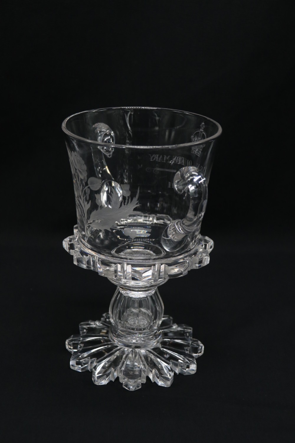 A clear glass commemorative goblet with two loop handles, wheel-engraved with text for the 1911 coronation of King George V and Queen Mary, supported by a faceted stem and starburst base. View IV