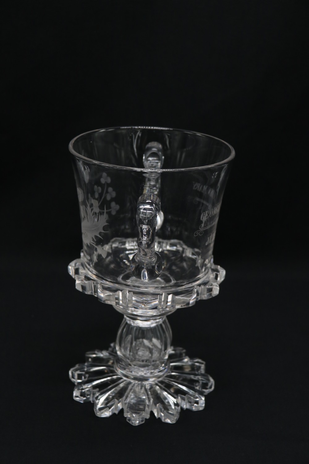 A clear glass commemorative goblet with two loop handles, wheel-engraved with text for the 1911 coronation of King George V and Queen Mary, supported by a faceted stem and starburst base. View IX