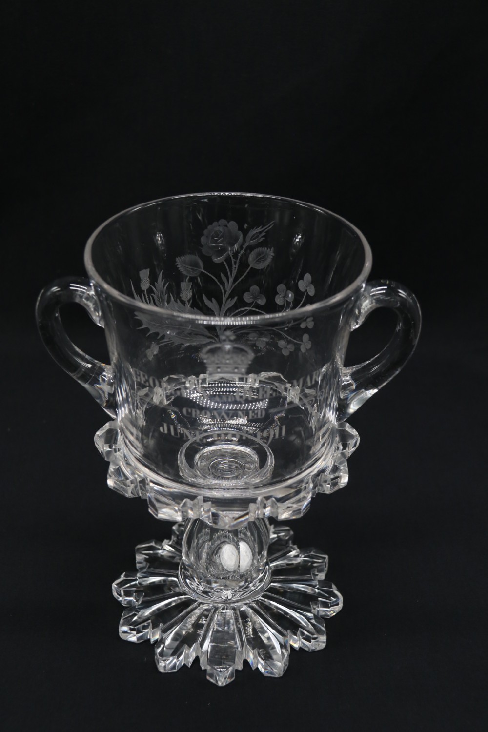 A clear glass commemorative goblet with two loop handles, wheel-engraved with text for the 1911 coronation of King George V and Queen Mary, supported by a faceted stem and starburst base. View XI
