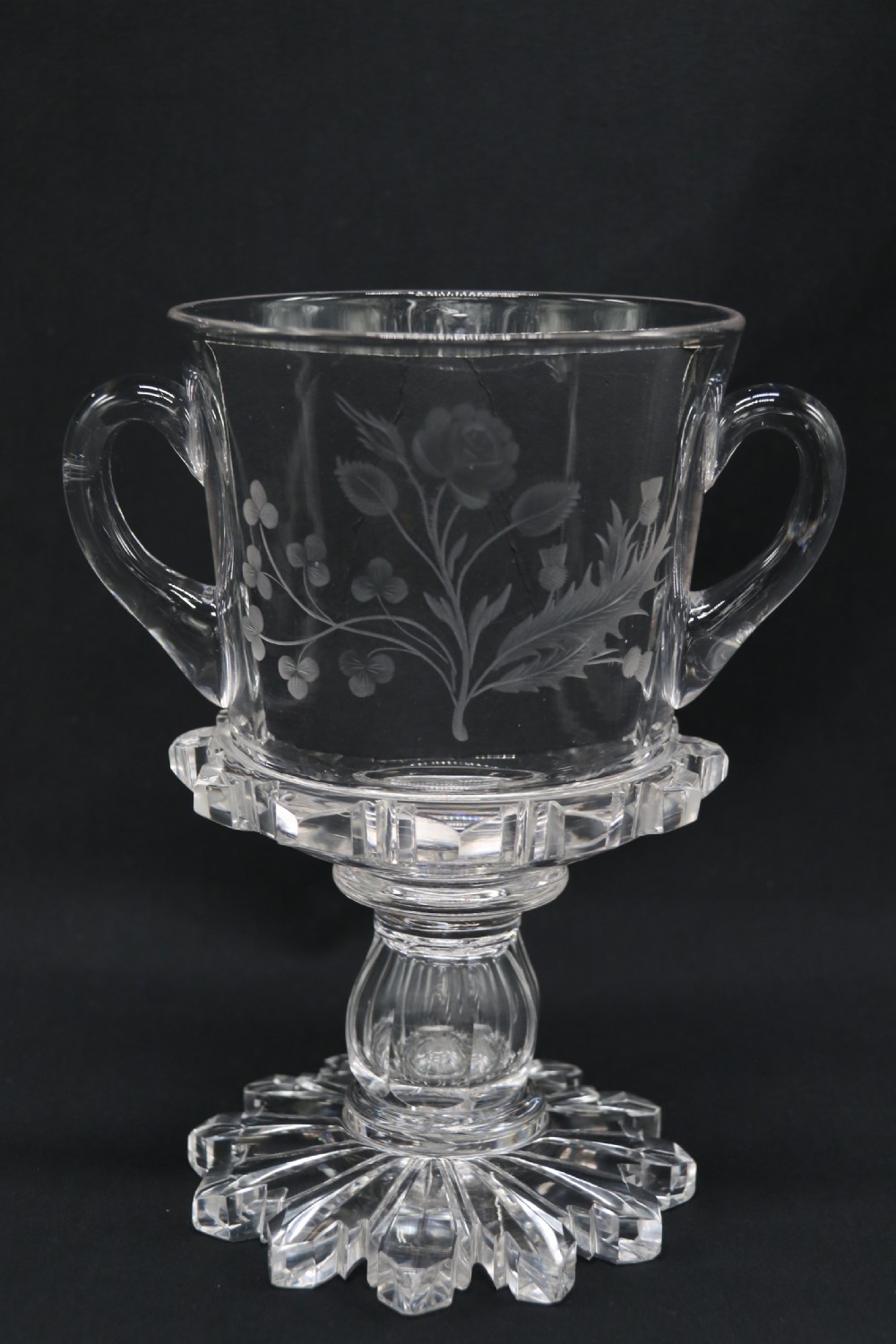 A clear glass commemorative goblet with two loop handles, wheel-engraved with text for the 1911 coronation of King George V and Queen Mary, supported by a faceted stem and starburst base. View XII