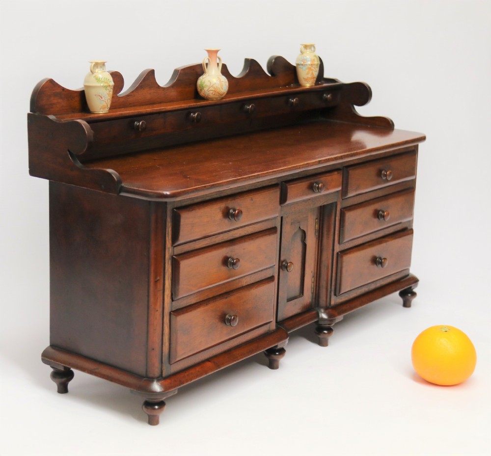 A nineteenth-century miniature mahogany dresser base featuring a shaped gallery back with pegs, multiple small drawers with turned knobs, and a central panelled cupboard door resting on turned feet.