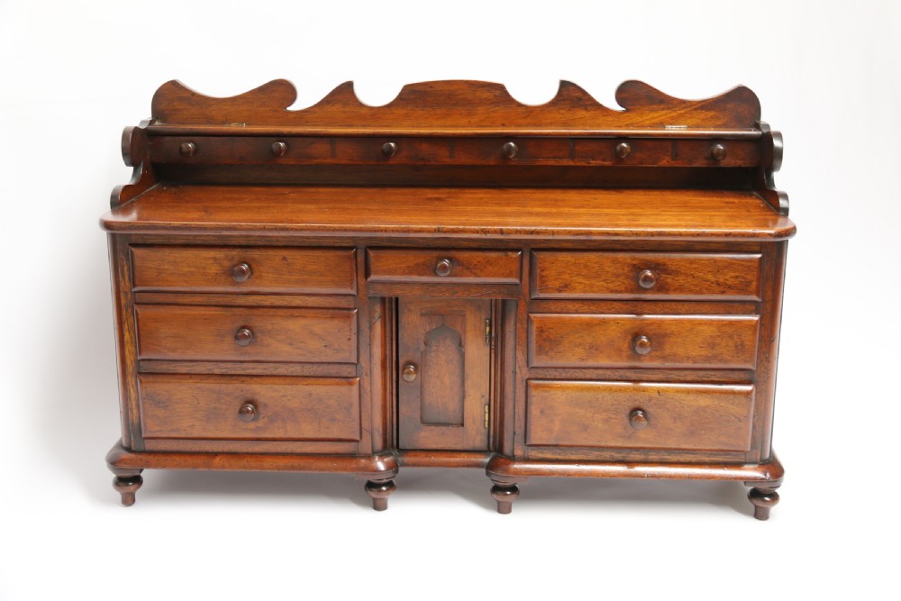 A nineteenth-century miniature mahogany dresser base featuring a shaped gallery back with pegs, multiple small drawers with turned knobs, and a central panelled cupboard door resting on turned feet. View II