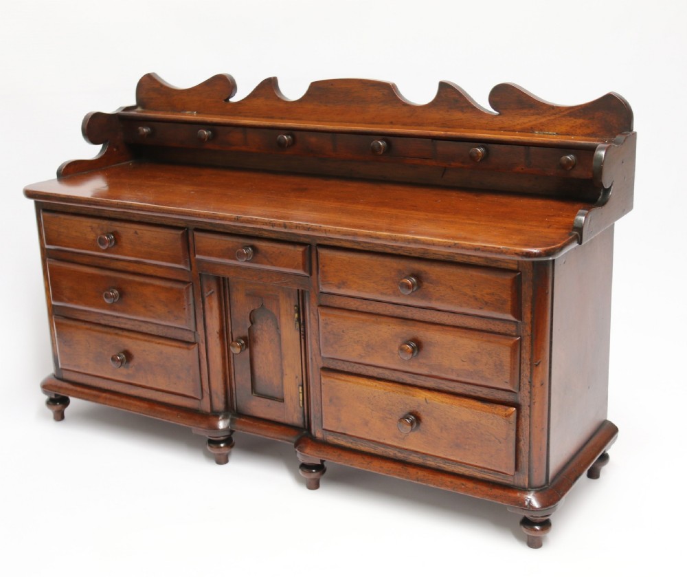 A nineteenth-century miniature mahogany dresser base featuring a shaped gallery back with pegs, multiple small drawers with turned knobs, and a central panelled cupboard door resting on turned feet. View III