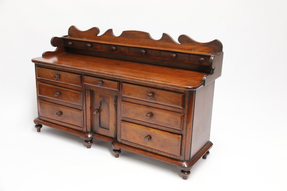 A nineteenth-century miniature mahogany dresser base featuring a shaped gallery back with pegs, multiple small drawers with turned knobs, and a central panelled cupboard door resting on turned feet. View IV