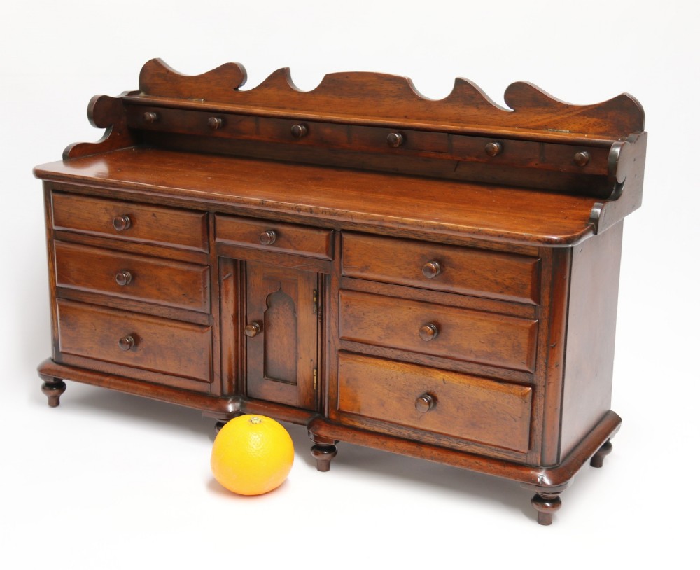 A nineteenth-century miniature mahogany dresser base featuring a shaped gallery back with pegs, multiple small drawers with turned knobs, and a central panelled cupboard door resting on turned feet. View V
