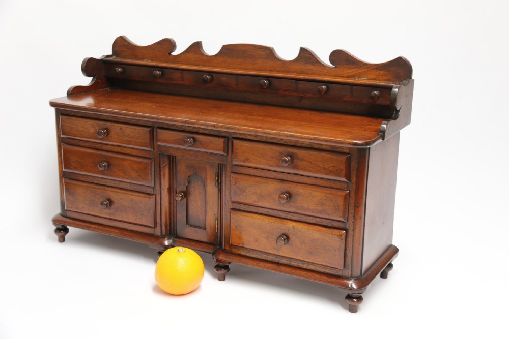 A nineteenth-century miniature mahogany dresser base featuring a shaped gallery back with pegs, multiple small drawers with turned knobs, and a central panelled cupboard door resting on turned feet. View VI
