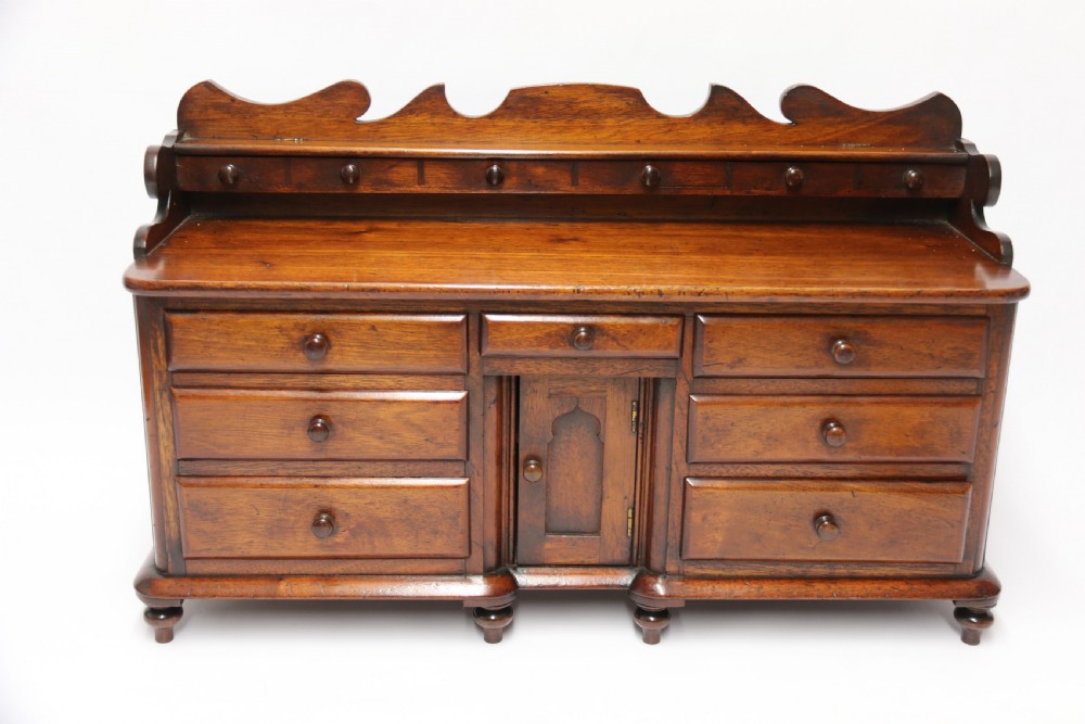 A nineteenth-century miniature mahogany dresser base featuring a shaped gallery back with pegs, multiple small drawers with turned knobs, and a central panelled cupboard door resting on turned feet. View VII