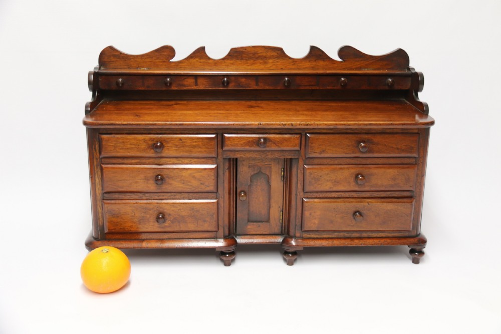 A nineteenth-century miniature mahogany dresser base featuring a shaped gallery back with pegs, multiple small drawers with turned knobs, and a central panelled cupboard door resting on turned feet. View VIII