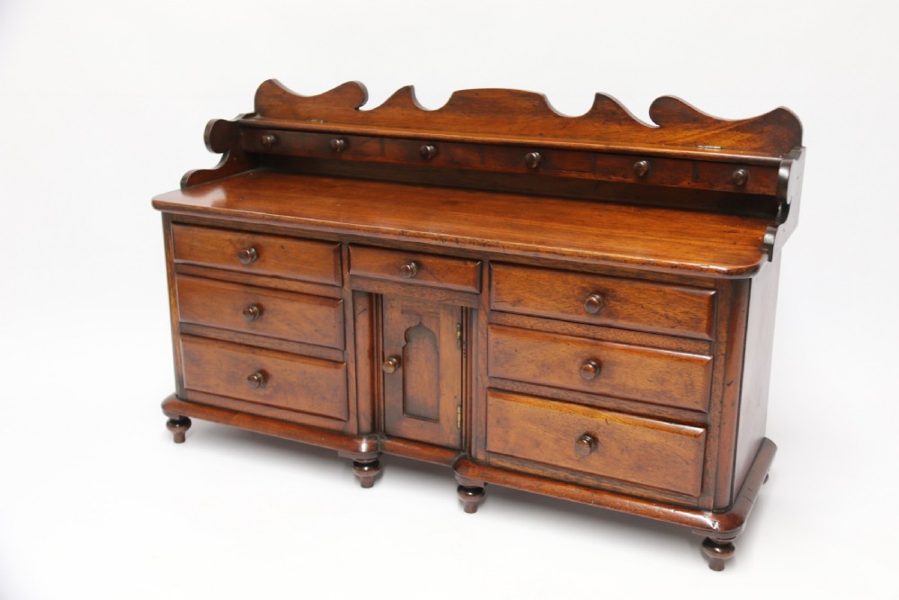 A nineteenth-century miniature mahogany dresser base featuring a shaped gallery back with pegs, multiple small drawers with turned knobs, and a central panelled cupboard door resting on turned feet. View IX