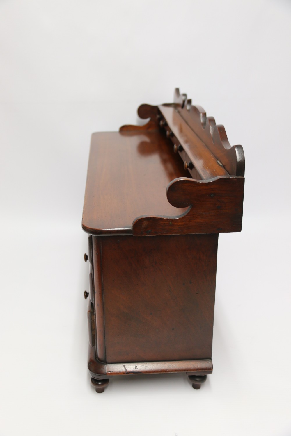 A nineteenth-century miniature mahogany dresser base featuring a shaped gallery back with pegs, multiple small drawers with turned knobs, and a central panelled cupboard door resting on turned feet. View X