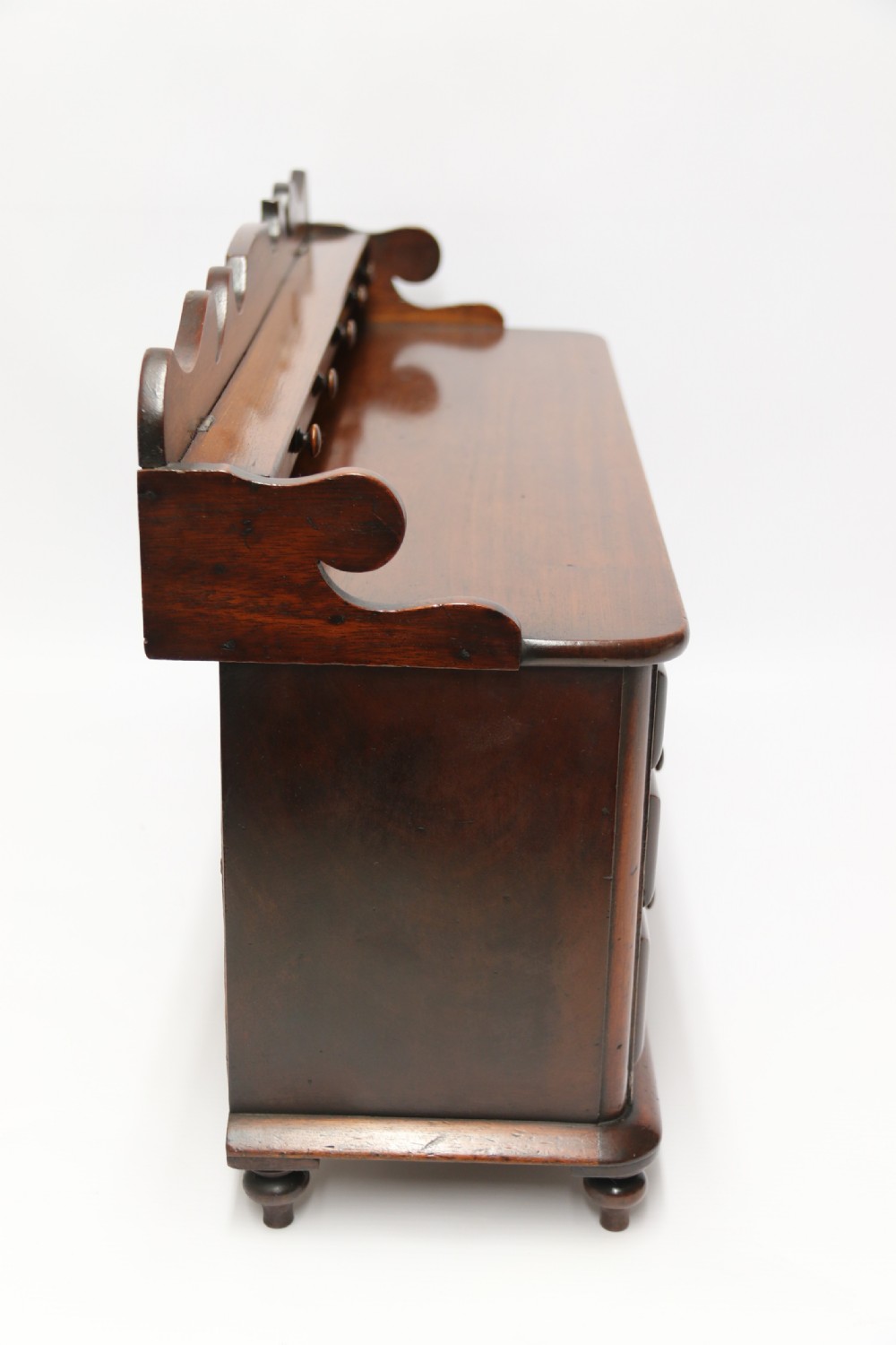 A nineteenth-century miniature mahogany dresser base featuring a shaped gallery back with pegs, multiple small drawers with turned knobs, and a central panelled cupboard door resting on turned feet. View XII