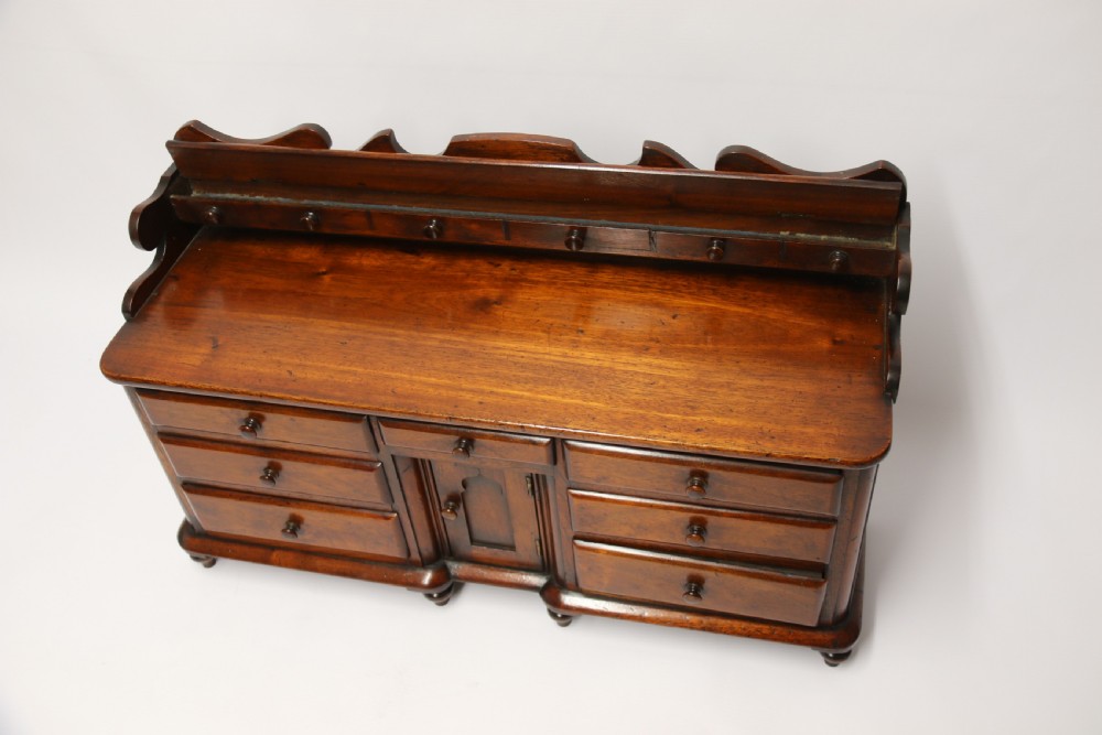 A nineteenth-century miniature mahogany dresser base featuring a shaped gallery back with pegs, multiple small drawers with turned knobs, and a central panelled cupboard door resting on turned feet. View XIII