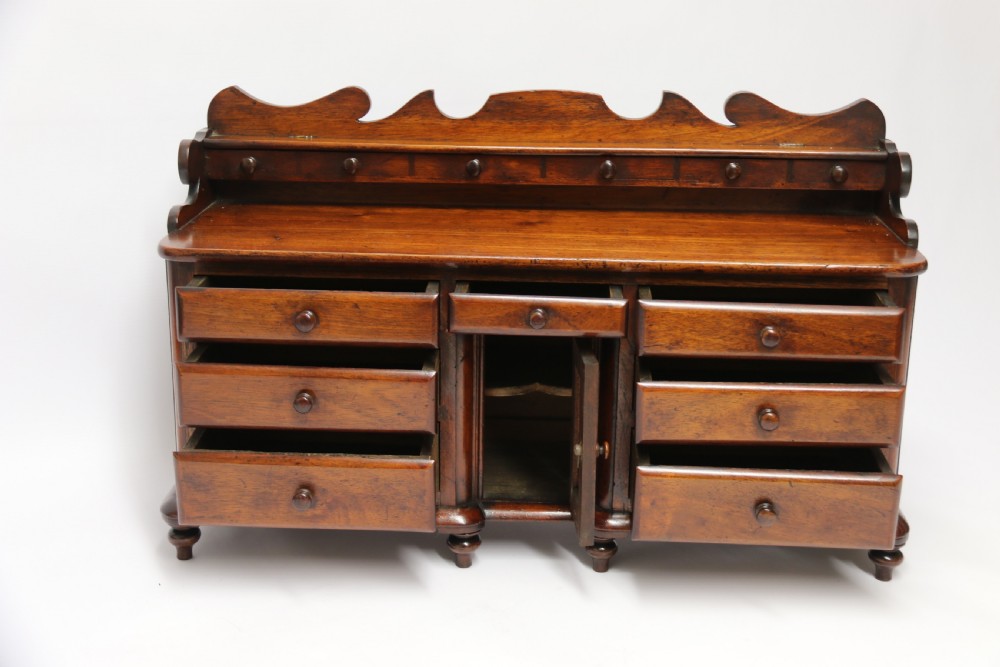 A nineteenth-century miniature mahogany dresser base featuring a shaped gallery back with pegs, multiple small drawers with turned knobs, and a central panelled cupboard door resting on turned feet. View XV