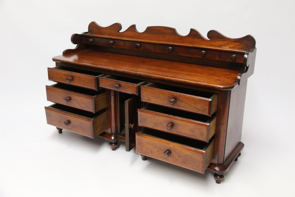 A nineteenth-century miniature mahogany dresser base featuring a shaped gallery back with pegs, multiple small drawers with turned knobs, and a central panelled cupboard door resting on turned feet. View XVI
