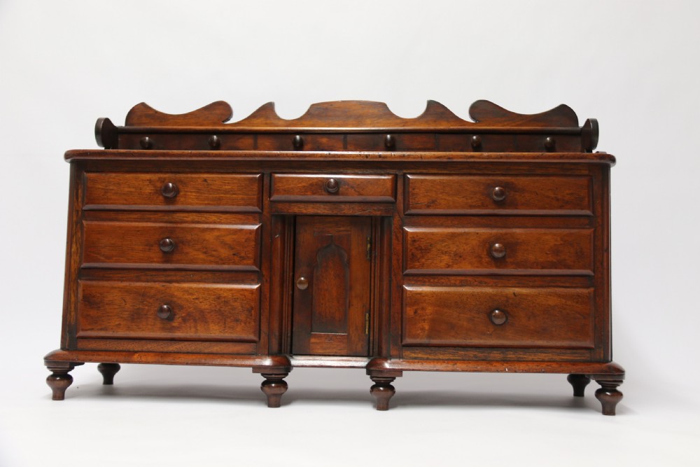 A nineteenth-century miniature mahogany dresser base featuring a shaped gallery back with pegs, multiple small drawers with turned knobs, and a central panelled cupboard door resting on turned feet. View XVII