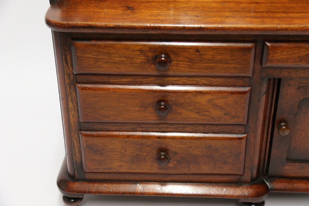 A nineteenth-century miniature mahogany dresser base featuring a shaped gallery back with pegs, multiple small drawers with turned knobs, and a central panelled cupboard door resting on turned feet. View XVIII