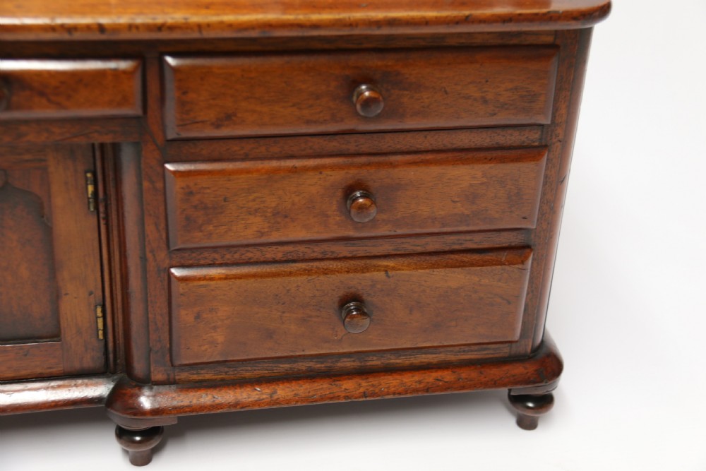 A nineteenth-century miniature mahogany dresser base featuring a shaped gallery back with pegs, multiple small drawers with turned knobs, and a central panelled cupboard door resting on turned feet. View XX