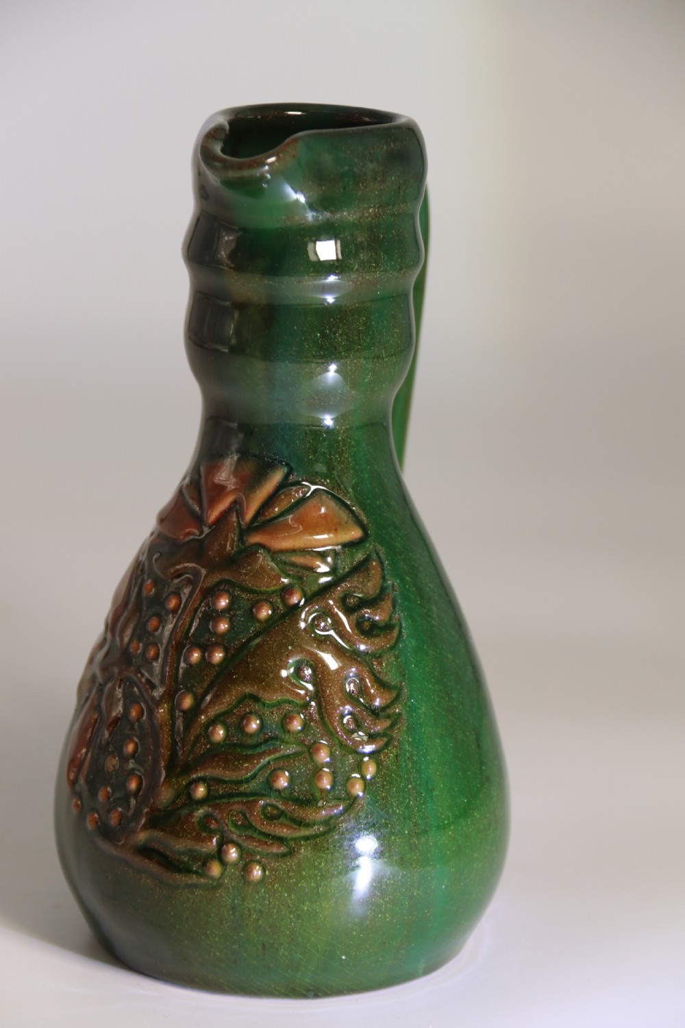 A green glazed ceramic jug featuring a flared spout, loop handle, and an embossed sunflower motif on the side. View V