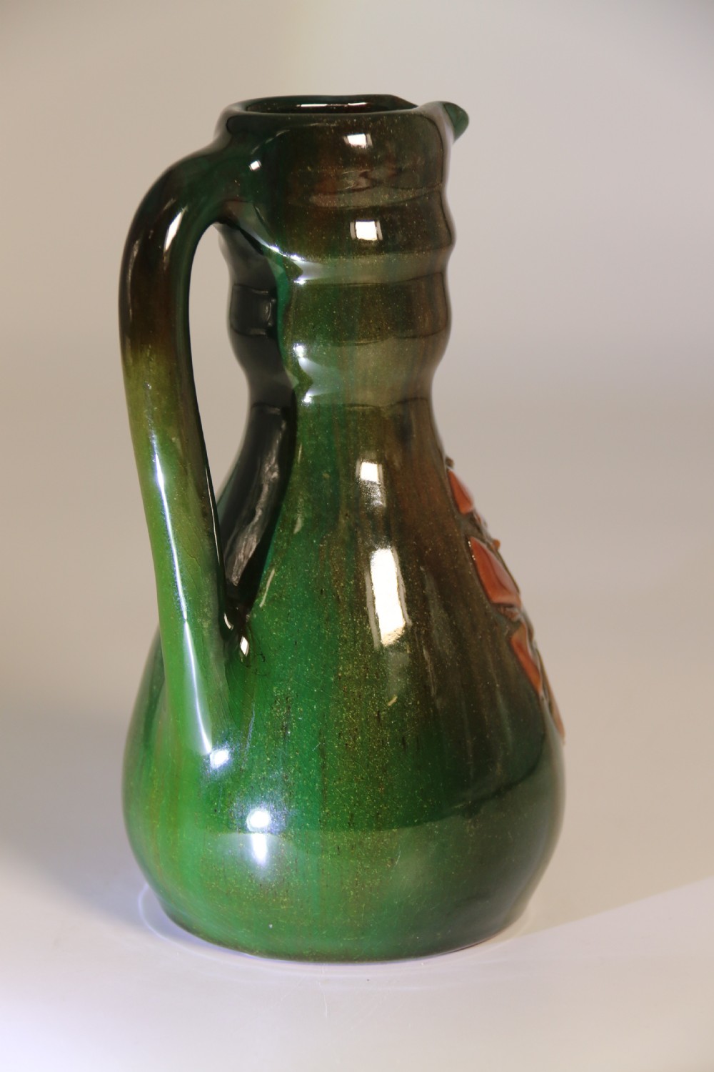 A green glazed ceramic jug featuring a flared spout, loop handle, and an embossed sunflower motif on the side. View VII